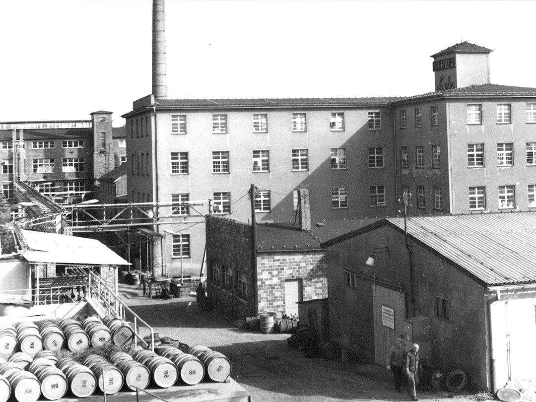 Historische Schwarz-Weiß-Aufnahme des OLI LACKE -Betriebshofs mit Produktionsgebäuden und Holzfässern im Vordergrund.
