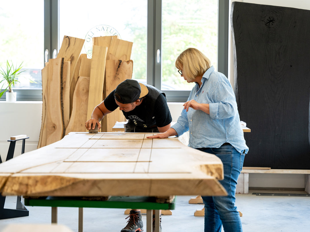 Zwei Menschen begutachten und bearbeiten gemeinsam die Oberfläche einer großen Holzplatte in einer Werkstatt.