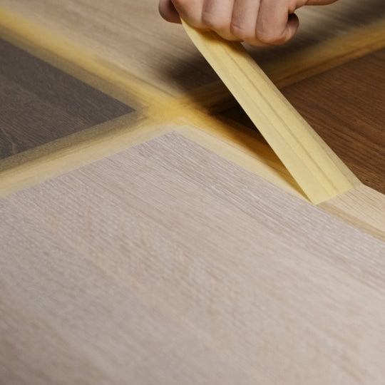 Eine Hand zieht Abklebeband zwischen unterschiedlich gefärbten Holzmusterflächen ab.
