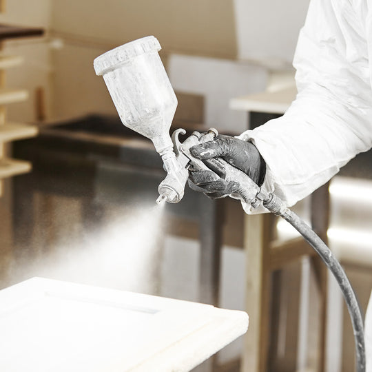 Verarbeiter mit schwarzen Handschuhen lackiert eine Holzoberfläche mit weißem Lack und einer Spritzpistole.